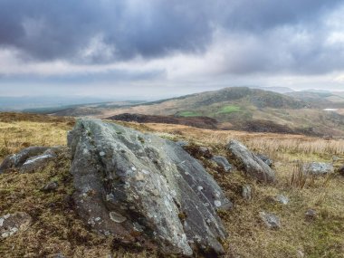 Kasvetli, tepelik bir manzara kendini gösteriyor. Gri bir gökyüzü ve dağınık kayalar görülebilir. Uzak tepeler kara bulutların altında beliriyor.