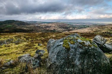 Çimenli bir tepenin üzerinde büyük, düzensiz bir kaya koleksiyonu duruyor. Arka plan bulutlarla dolu bir vadiyi ve gökyüzünü gösteriyor..