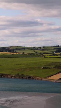 Yumuşak bulutların altında yemyeşil alanlar ve kırsal alanda sakin bir koy manzarası.
