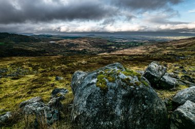 Değişen boyutlarda koyu gri kayalar yamaçta yığılmıştır. Geniş, düz bir vadi bulutlu bir gökyüzünün altında tepenin ötesine uzanır..