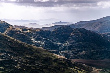 Görüntü dağlık bir bölgeyi gösteriyor. Karanlık, engebeli arazi, daha aydınlık ve uzak tepelerle çelişen ön planda hüküm sürüyor..