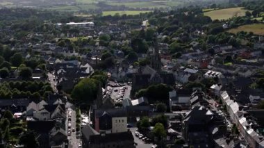 Video bir kasabanın çatılarını gösteriyor, çoğu gri, bol yeşillikle çevrili, yüksek bir kilise kulesi görülebiliyor..