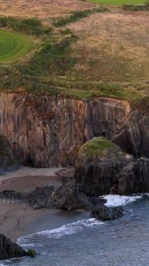 Çimenli bir yamaç, karanlık bir kayalık kıyı şeridi boyunca çeşitli deniz mağaralarına ve körfezlere bakar..