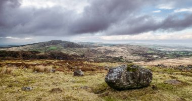 Önünde büyük kayalar, arkasında tepeler ve bulutlu gökyüzü olan otlak bir alan..