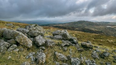 Tepenin yamacında sergilenen ve yeşilliklerle kaplı tepeleri olan bir manzara manzarası. Gökyüzü bulutlu, arazide dramatik bir atmosfer yaratıyor..