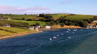 Beyaz evler ve küçük tekneler korunan bir bölmeyi işgal ediyor. Sahne huzurlu. Sakin suları çevreleyen yeşil tepeler. Güneşli bir gün.