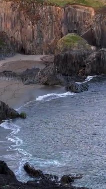 Video, kumlu sahili olan tenha bir koy gösteriyor. Koyu gri kayalar kara ve deniz arasında doğal bir bariyer oluşturur. Bitkiler kayalıkların tepelerini kaplar..