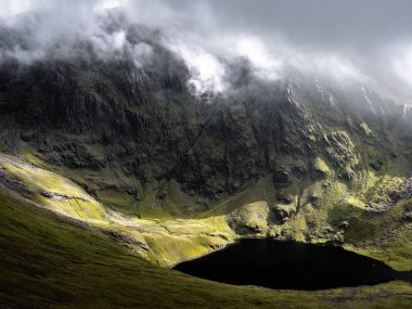 İrlanda 'da dingin, karanlık bir gölü çevreleyen engebeli dağların nefes kesici manzarası. Manzara yosunlu yeşilliklerle ve yukarıdaki bulutlardan gelen gizli gölgelerle dolu..