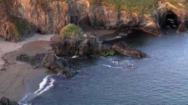 Dar bir kumsalın olduğu sahil manzarası. Büyük, karanlık kayalar denize dökülüp engebeli bir kıyı şeridi oluşturuyor. Su nispeten sakin..