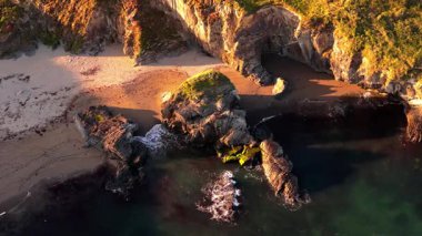 Kayalık bir sahil şeridinin yüksek açılı görüntüsü. Bir plaj kısmen görülebilir. Okyanus suyu karanlık görünüyor. Gölgeler kumlara dökülür.