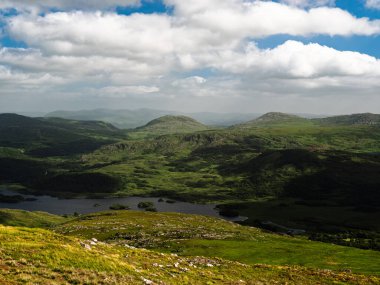Görüntü birkaç tepe ve dağı gösteriyor. Ön planda bir göl var. Gökyüzü açık bulutlu ve mavi lekeli..