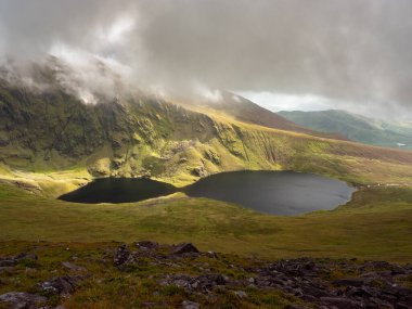 County Kerry 'deki Coomloughra Lough karanlık ve yükselen bulutların altında büyüleyici, bereketli bir manzara sunuyor. Sakin göller etrafındaki dağları yansıtıyor ve nefes kesici bir manzara yaratıyor..