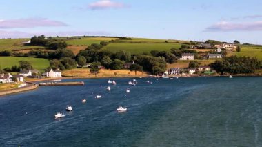 Sakin bir körfez sahnesi. Rıhtımda beyaz evler görülebiliyor ve yakınlarda birçok küçük tekne bulunuyor. Arka planda yeşil tepeler var..