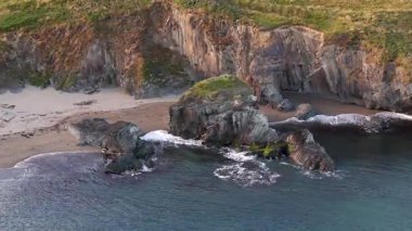 Video, dramatik bir sahil şeridi sergiliyor. Dik kayalıklar, koyu renkli kayalar ve küçük kumlu bir plaj göze çarpan özelliklerdir. Su koyu mavi-gri..