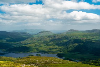 Bir vadiyi resmeden manzara fotoğrafı. Özellikler arasında bir göl, tepeler ve ağaçlar yer alıyor. Gökyüzü güneş ve bulutların karışımıdır..