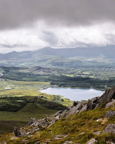 Huzurlu bir manzaranın yüksek açılı görüntüsü. Yuvarlanan tepeler ve vadiler arasında çeşitli bitki örtüleriyle çevrili bir göl görülebilir. Gri kayalar ön planda..