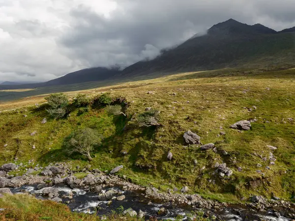 Gaddagh Nehri, bulutlu bir gökyüzü altında yuvarlanan tepeler ve dramatik dağlarla çevrili İrlanda 'nın yemyeşil manzarası boyunca yavaşça akar ve sakin bir atmosfer yaratır..