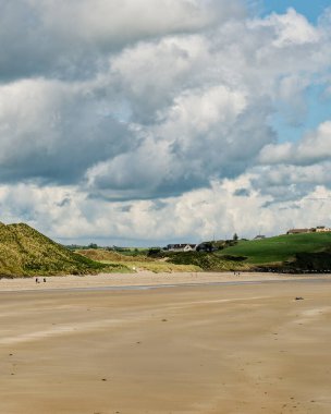 West Cork, 25 Haziran 2023. Huzurlu sahil manzarası. Altın kum yemyeşil tepeler ve uzakta birkaç evle buluşur. İnsanlar parlak ama bulutlu bir gökyüzünün altında kıyı boyunca yürür..
