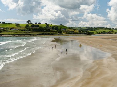 West Cork, 25 Haziran 2023. Huzurlu sahil manzarası. Nazik dalgalar, parçalı bulutlu bir gökyüzünün altında güneşli bir günün tadını çıkaran figürlerle dolu kumlu bir sahili okşar. Yuvarlanan yeşil tepeler resim gibi bir zemin oluşturuyor..