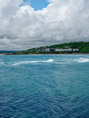 İnchydoney Adası 'nın üzerinde bulutlar toplanırken dalgalar yavaşça kıyıya doğru akar ve sakin manzarasını aydınlatır. Parlak binalar canlı yeşilliklerin arasına yerleşmiş, barışçıl bir geri çekilme yaratıyorlar..