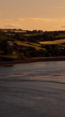 Low Tide Nehri. Alçak gelgit, geniş bir nehir kıyısındaki kumları ortaya çıkarır. Etrafı yavaşça yuvarlanan yeşil tepeler ve dağınık evlerle çevrilidir..