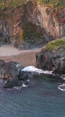 Yüksek irtifa videosu engebeli, kayalık bir kıyı şeridini gösteriyor. Küçük bir kumlu koy karanlık, katmanlı uçurumlarla çevrilidir. Küçük dalgalar kıyıya ulaşır.