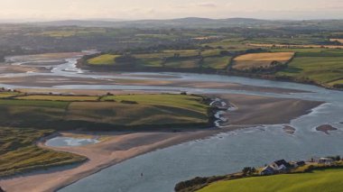 Sarmal nehir, gün batımında dingin İrlanda kırsalındaki canlı yeşil tarlalarda akar ve manzaranın doğal güzelliğini ve sükunetini ön plana çıkarır..