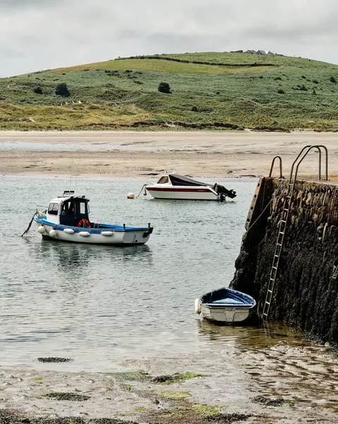 West Cork, 18 Haziran 2023. Tekneler İrlanda 'daki Middle Ring Bay Sahili' nin sığ sularında dinleniyor. Arkaplanda yemyeşil tepeler yükseliyor bulutlu bir gökyüzünün altında, huzurlu bir kıyı manzarası yaratıyor..