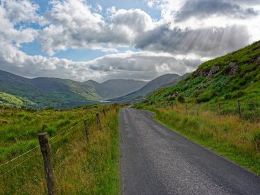 Sarmal bir yol, İrlanda, BlackValley 'in güzel yeşil manzarasını kesiyor. Görkemli dağlarla çevrili, dramatik bulutlarla dolu bir gökyüzünün altında..