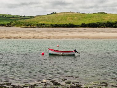 Küçük bir tekne İrlanda 'daki Middle Ring Bay Sahili' nin sığ sularında huzur içinde yatıyor. yemyeşil tepeler bulutlu bir gökyüzünün altındaki arkaplanı çerçeveleyerek sakin bir atmosfer yaratıyor..