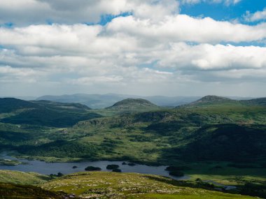 Farklı bölgeleri gösteren yüksek açılı bir çekim. Yeşil tepeler, karanlık ormanlar ve bir su kütlesi. Gökyüzü çoğunlukla bulutlu.