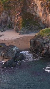 Bu bir kıyı bölgesinin genel görünümü. Karanlık, katmanlı kayalar ve dik bir uçurum yüzeyi ile kısmen çevrili küçük kumlu bir sahili vurgular. Su karanlık..