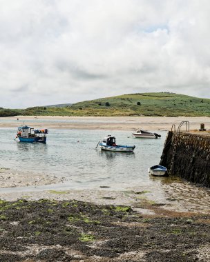 West Cork, 18 Haziran 2023. Botlar İrlanda 'daki Middle Ring Bay Sahili' nin sığ sularında huzur içinde dinleniyor. Arkaplanda yemyeşil tepeler yükseliyor bulutlu bir gökyüzünün altında, sakin bir kıyı manzarası yaratıyor.