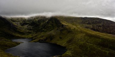 Coomloughra Lough, tepede yemyeşil tepeler ve dramatik bulutlarla çevrili karanlık suları sergiliyor. Huzurlu ortam, doğanın harikalarını keşfetmeye ve takdir etmeye davet ediyor..