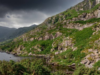 Huzurlu bir nehre bakan görüntü, yeşillikle örtülü sarp kayalık tepeleri yakalıyor. Üzerimizde kara bulutlar beliriyor, huzurlu ortama dram katıyor..