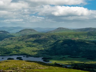 Yeşil bir manzaranın geniş bir görüntüsü. Yuvarlanan tepeler, bir göl ve parçalı bulutlu bir gökyüzü. Uzak dağlar görülebilir..