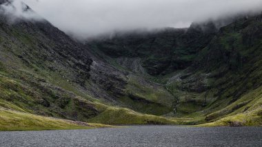 Coomloughra Lough, İrlanda 'daki sarp, yeşil kaplı dağların eteğinde huzur içinde yatıyor. Bir bulut örtüsü üzerinde süzülüyor, bu doğal ortamın huzur veren güzelliğini arttırıyor..