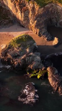 Küçük bir kumsal içeren kıyı manzarası. Karanlık kayalar okyanusa dalıyor. Bitki örtüsü yukarıdaki kayalıkları kaplıyor. Dalgalar görünür..