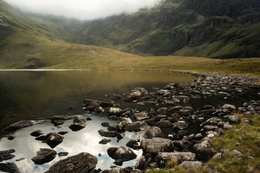 Etrafı kayalık kıyı şeridi ve sisli yeşil tepelerle çevrili sakin bulutlu gökyüzünü yansıtan dingin dağ gölü..