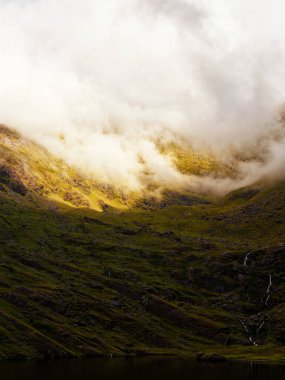 Çimenli bir yamaç Lough Callee 'nin karanlık sularına doğru iniyor. Tepenin tepesinde bulutlar asılı, İrlanda 'da sakin ve heyecan verici bir manzara yaratıyor..