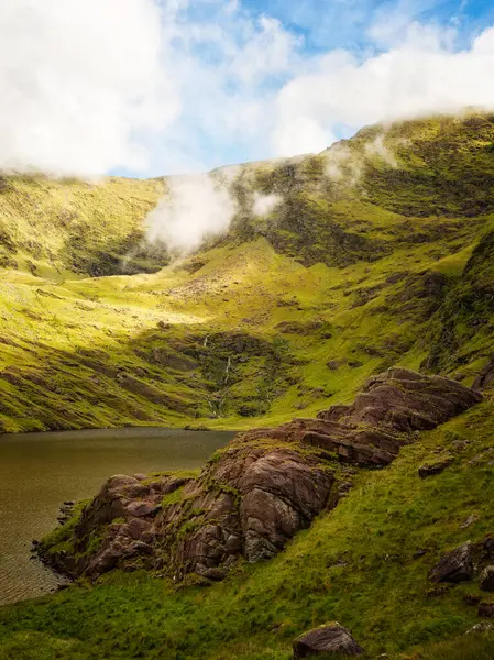 Yeşil yamaçlar Lough Callee 'ye gidiyor. Yukarıdaki mavi gökyüzü ile tepelerin üst kısımlarında sis asılı. Göl kenarında kayalar görülebilir..
