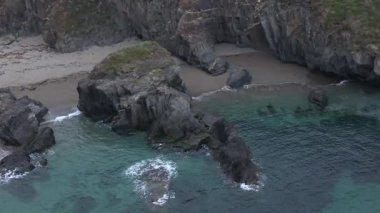 Dalgalar kayalık kıyıya hafifçe çarpar. Uçurumlar ve kaya oluşumları arasında küçük, tenha bir sahil vardır. Turkuaz su, koyu kayaları çevrelemiş..