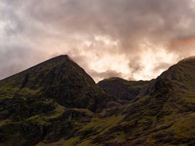 Carrauntoohil, İrlanda 'nın en yüksek zirvesi, alacakaranlık yaklaşırken dönen bulutların kulesine karşı. Canlı manzara yemyeşil tepeleri ve kayalık çıkıntıları sergiliyor..