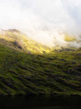 Çimenli bir yamaç Lough Callee 'nin karanlık sularına doğru iniyor. Tepenin tepesinde bulutlar süzülüyor, İrlanda 'da sakin ve dramatik bir manzara yaratıyor..