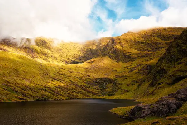 İrlanda 'nın Kerry ilçesindeki yeşil dağların arasına yerleşmiş Lough Callee' nin gündüz gösterileri. Güneş ışığı dağları aydınlatır. Hafif bulutlar tepeleri kısmen gizler..