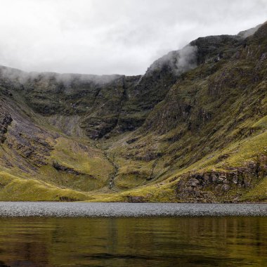 Dramatik dağlık arazi, sakin bir gölün sisli bir dağ sırasını yansıttığı nefes kesici bir manzara sunar..