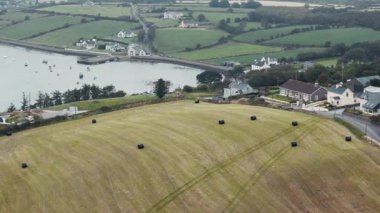 Güzel bir İrlanda manzarası, limanın yanındaki bir tarlada saman balyalarını, kıyı şeridine yayılmış evler ve tekneleri barındırır..