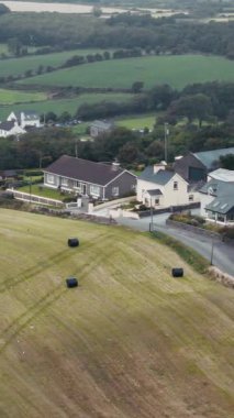Güzel bir İrlanda manzarası gösteriliyor. Limanın yanındaki tarlada saman balyalarını gösteriyor. Kıyı şeridi boyunca evler ve tekneler de var..