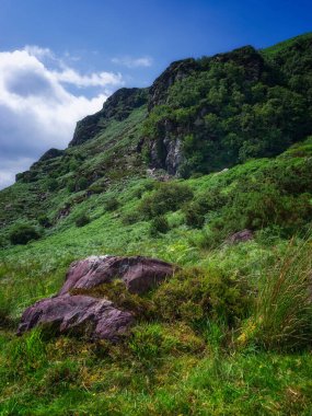 İrlanda 'nın Kerry ilçesinde açık, parçalı bulutlu bir günde, yemyeşil ve kayalarla kaplı sert dağ yüzeyi..