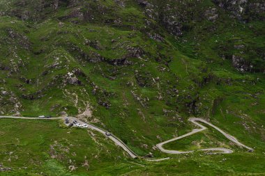 Dunloe, İrlanda, 1 Temmuz 2023. Araçlar, Dunloe geçidinde dolambaçlı bir yol boyunca seyahat ederler. Yol yeşil bitki örtüsüyle kaplı dik bir yamaca tırmanıyor, açıkta kayalar ve kayalıklar var..